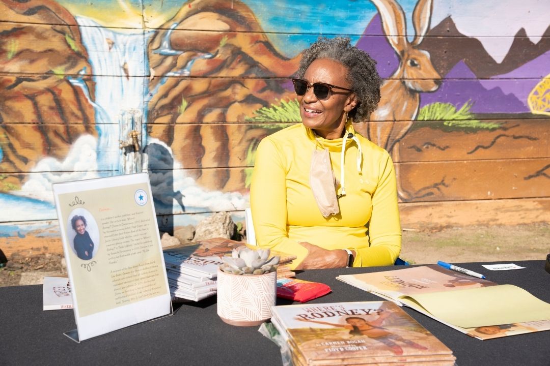 A Black femme in a yellow cardigan and sunglasses sits behind a table displaying the book she wrote. She is looking to the left and smiling at someone off camera. Behind her is a semi-realistic mural depicting a waterfall in a desert.