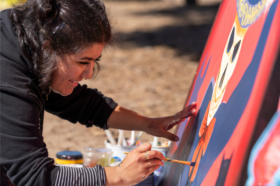 A smiling woman grins while painting a large mural in bright colors. She's bracing one paint-covered hand against the mural board while painting with the other.