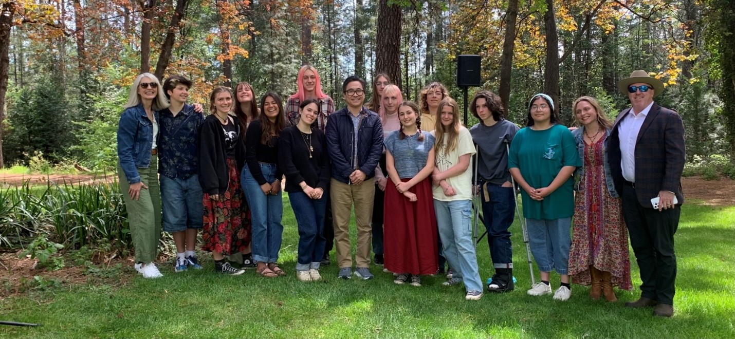 17 youths and adults stand smiling in shady grass with a forest backdrop. On the far left is Kirsten Casey, a woman with long white hair and dark sunglasses. In the center is Lee Herrick, a man with short dark hair and glasses. He is the 10th California Poet Laureate, and the first Asian American to serve in the role. On the right is Scott Green, a man in a park ranger hat, dark sunglasses, and a black suit.