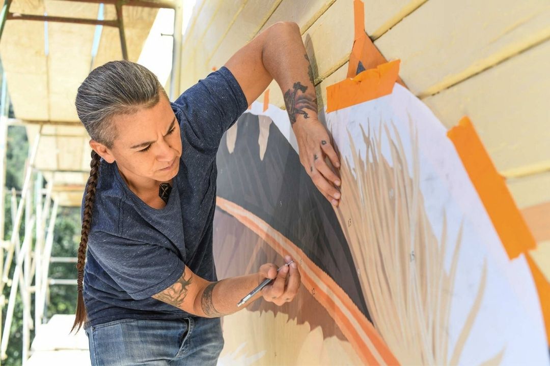 An artist carefully outlining a mural on a wall. The artist has long, dark hair braided down their back and is wearing a casual blue shirt and jeans. They are focused on the task, holding a tool in one hand while using the other hand to stabilize a paper template taped to the wall. The wall is in the process of being painted, with various shades of brown, beige, and orange visible on the mural. Scaffolding is present in the background.