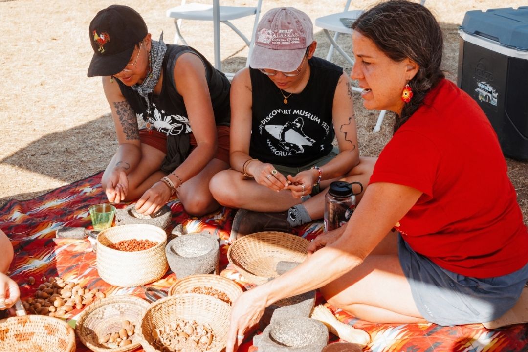 Three individuals are seated on a colorful red and orange textile spread on the ground. They are gathered around several woven baskets filled with acorns and seeds, along with stone mortars and pestles. A person in a red shirt on the right appears to be instructing the other two, who are focused on the materials in their hands. In the background, white folding chairs and a cooler.