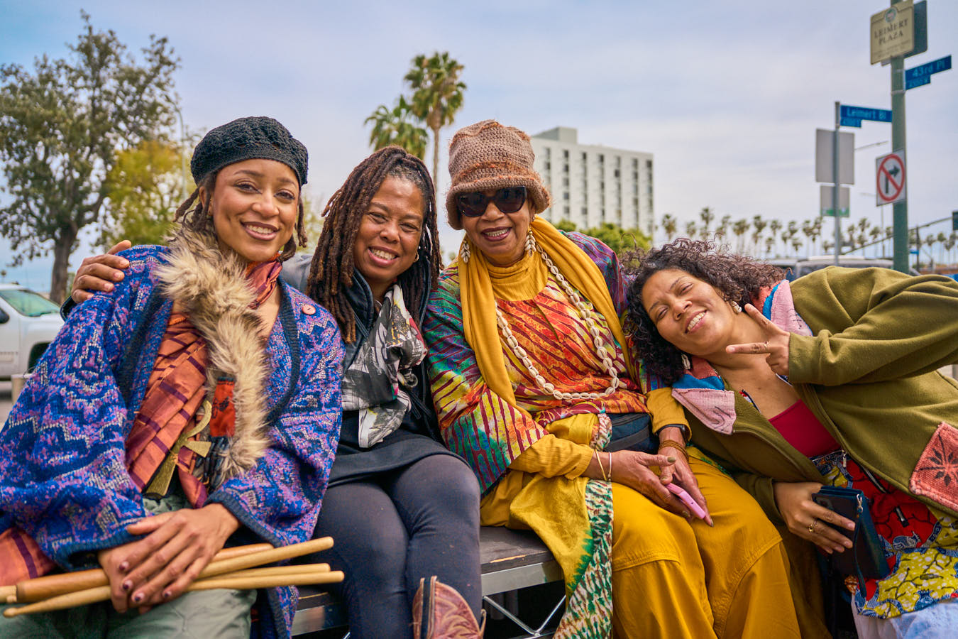 A group of four Black women sit on a bench, dressed in colorful, patterned layers, including knit hats, scarves, and jewelry. The woman on the far left holds a bundle of drumsticks. The background shows palm trees, a city building, and a bright, overcast sky.