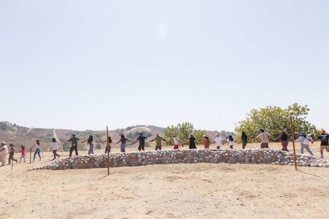 A group of people stands in a long line, holding hands. They are standing behind a long, low wall made of smooth river stones, with brown hills in the background.